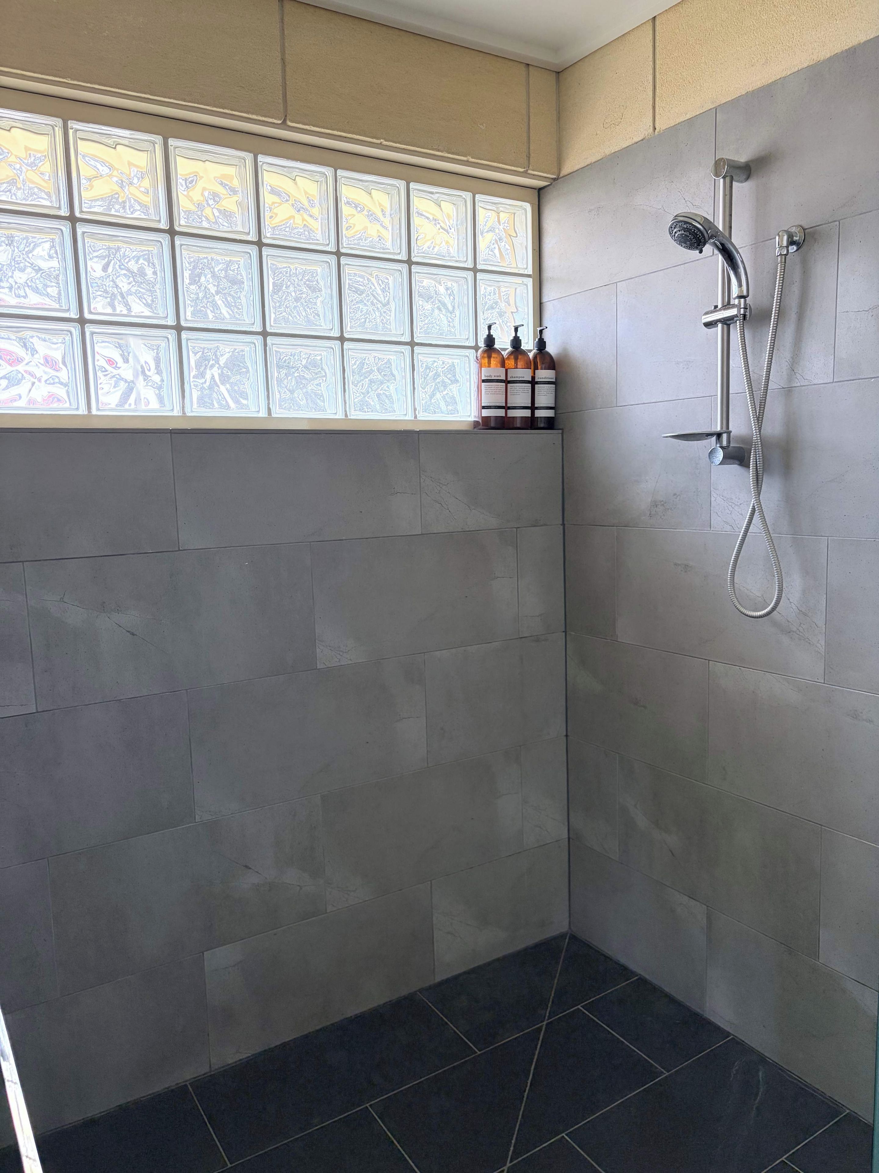 A bathroom with grey tiled walls, a shower with a handheld showerhead, and a window with decorative glass panels.