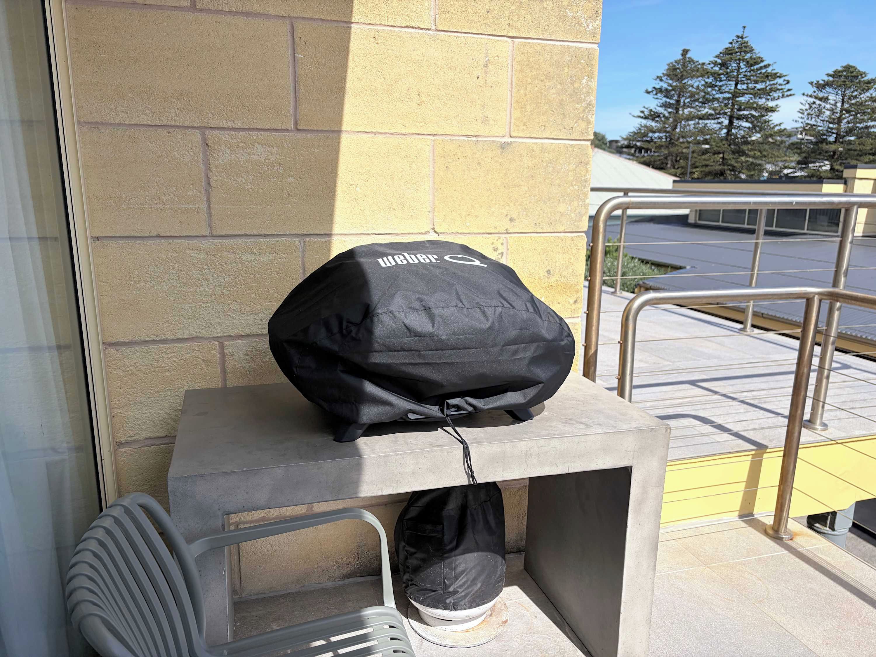 Una parrilla Weber negra sobre una mesa de concreto frente a un edificio con pared amarilla y barandilla de metal.