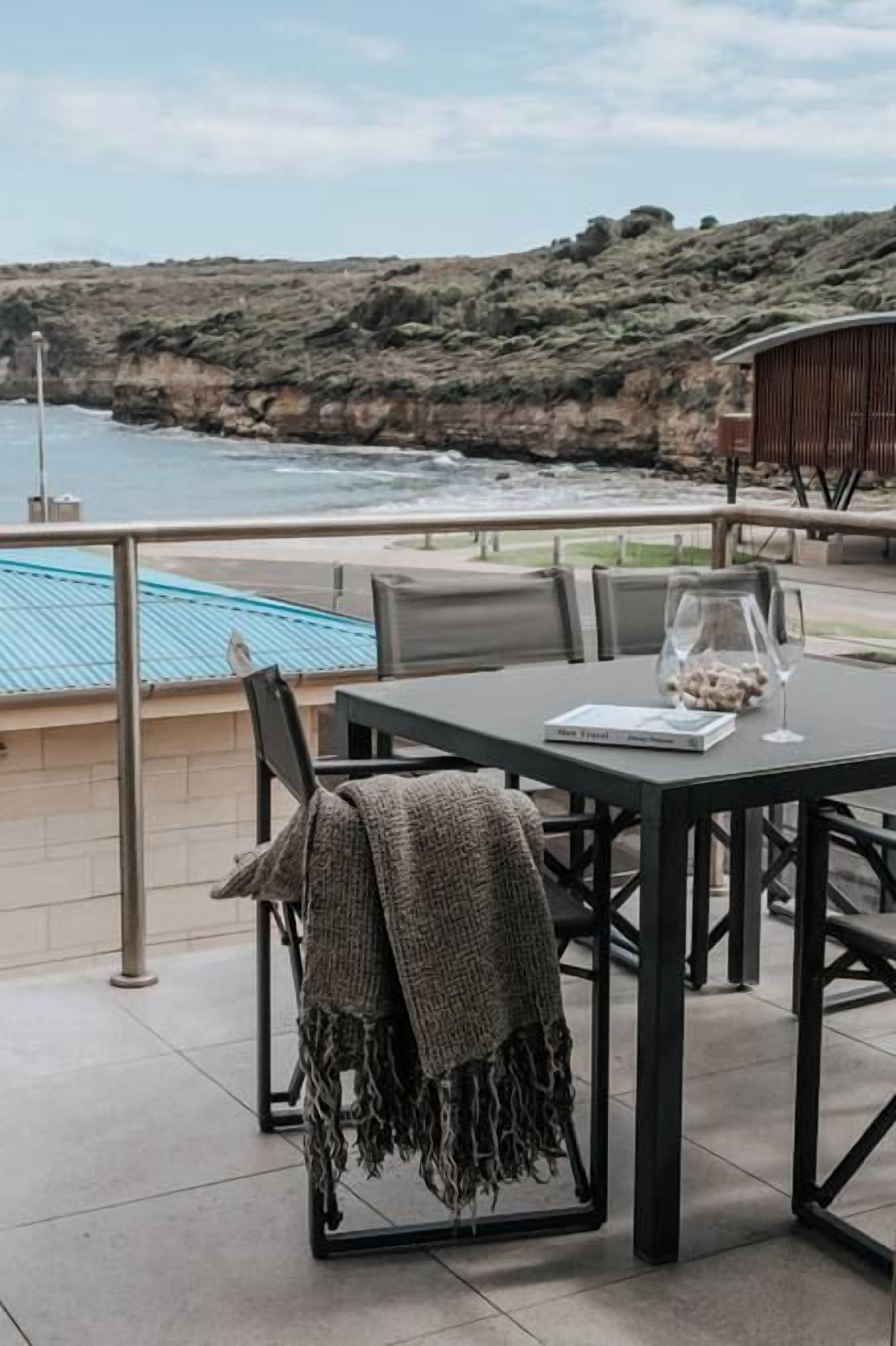 Une terrasse avec une table et des chaises, offrant une vue sur une côte rocheuse et une étendue d’eau.
