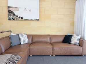 Ein braunes Ledersofa mit gemusterten Kissen vor einer Ziegelwand, darüber ein gerahmtes Bild einer Küstenlandschaft und rechts ein weißer Vorhang.