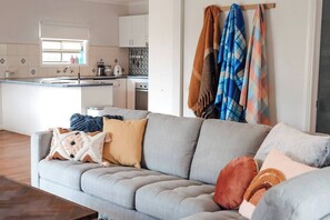 Un salon moderne avec un canapé gris, une cuisine en arrière-plan et une fenêtre laissant entrer la lumière naturelle.