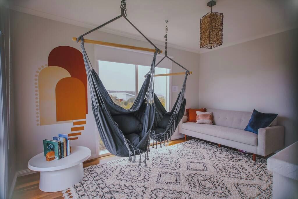 A modern living room with a hanging hammock chair, a white sofa, a patterned rug, and a minimalist wall design.
