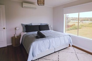 A bedroom with a bed, a bedside lamp, a window with a view, and an air conditioning unit.
