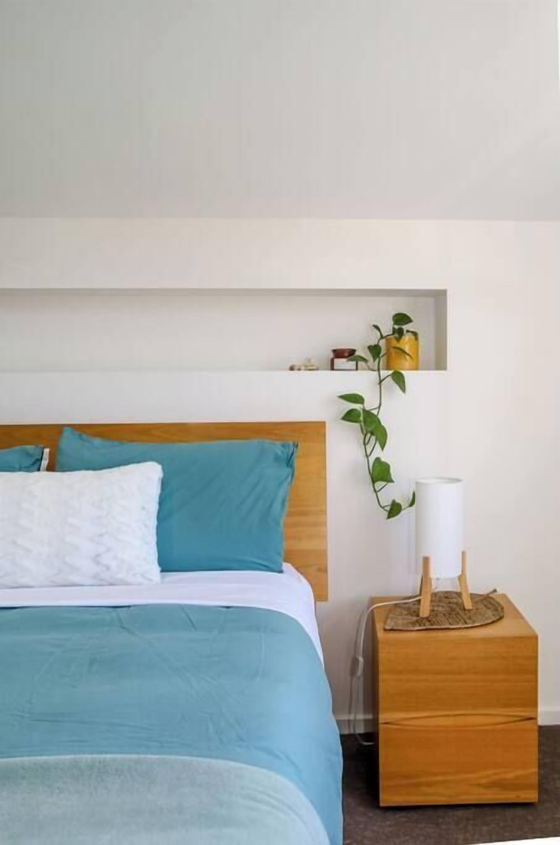 A bed with a wooden headboard, a bedside table with a lamp, and a shelf with a plant.