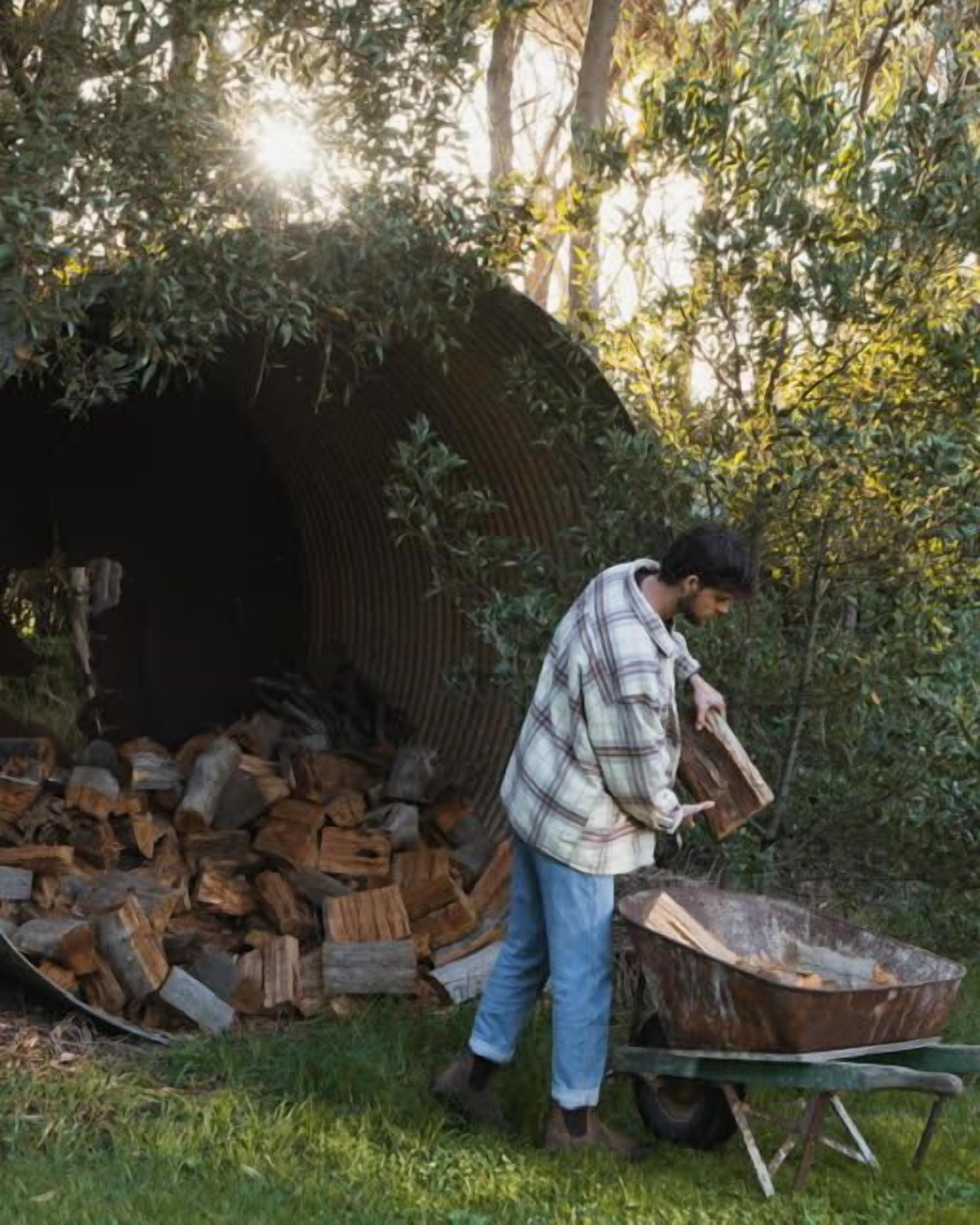 A person loading firewood into a wheelbarrow near a pile of logs and a tunnel-like structure.