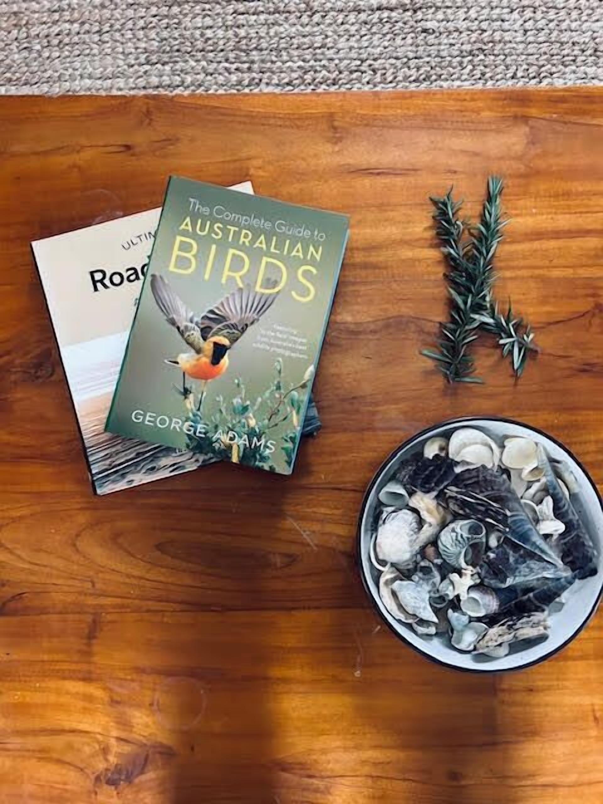 En bog med titlen 'The Complete Guide to Australian Birds' af George Adams på et træbord, ved siden af en skål med sneglehuse.