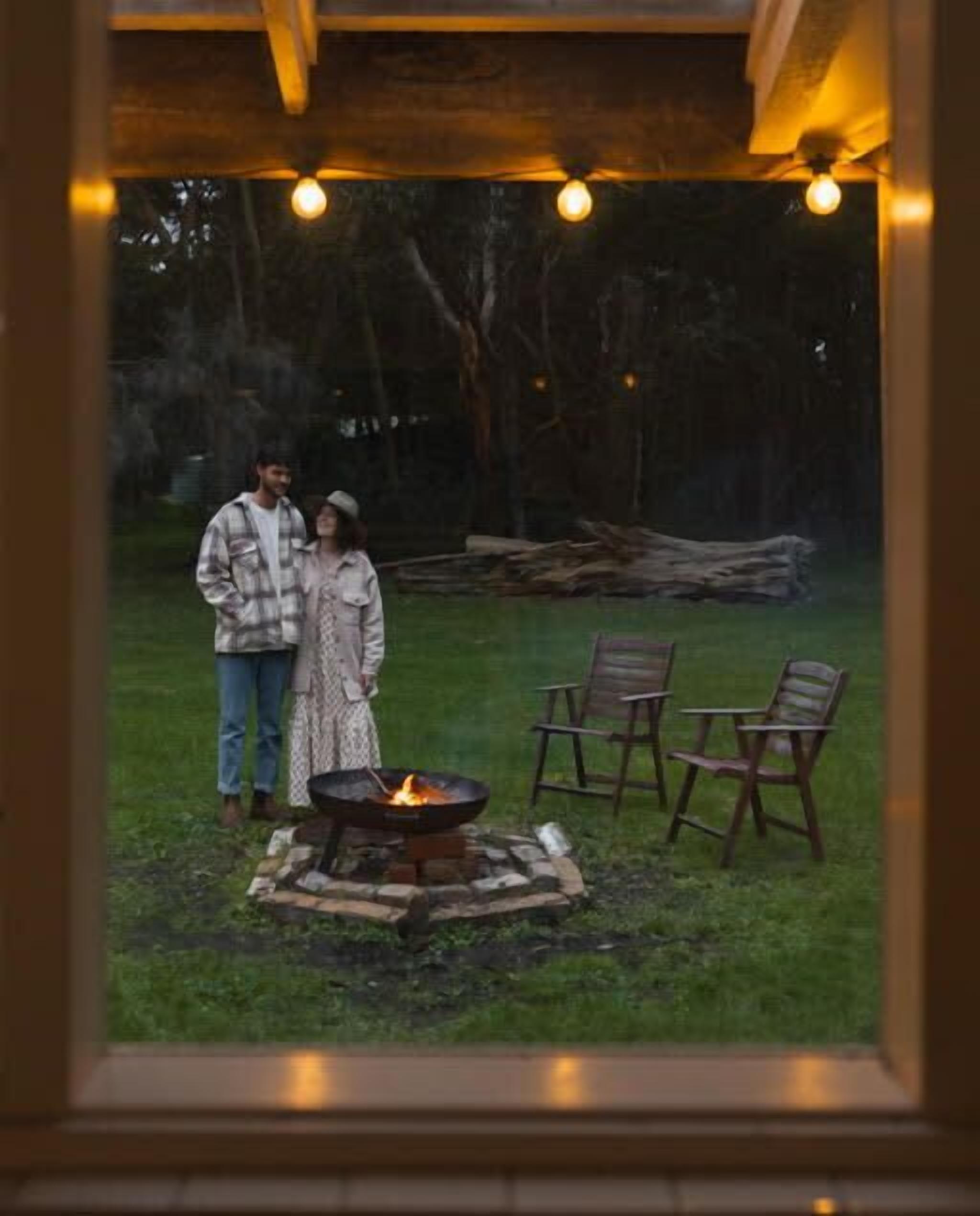 A couple standing by a fire pit with chairs and a wooden structure in a grassy outdoor area.
