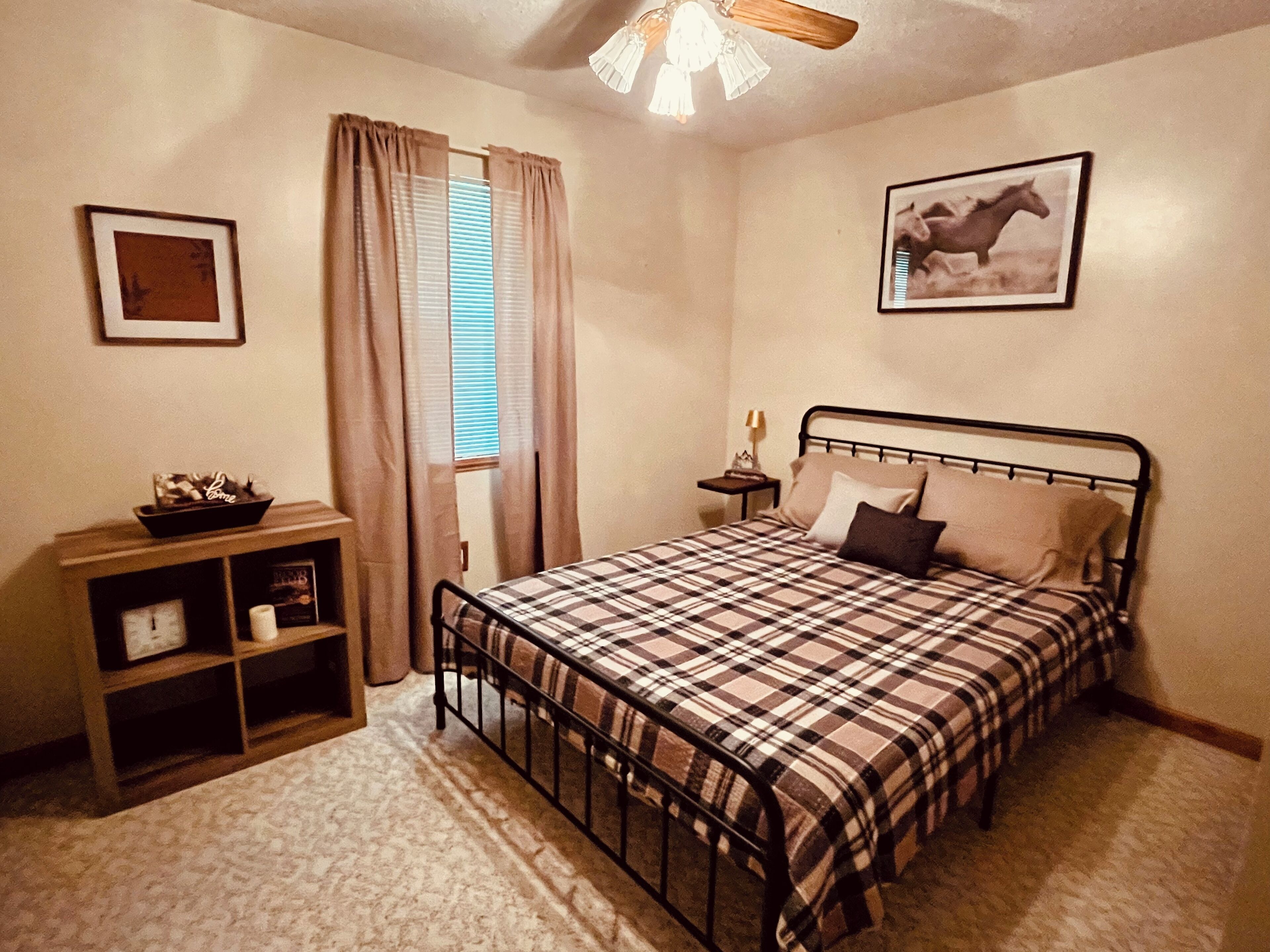 Un dormitorio con cama, mesita de noche, estante, ventana con cortinas, ventilador de techo y un cuadro enmarcado en la pared.
