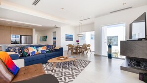 A modern living room with a sofa, coffee table, and fireplace. There is a dining area with chairs and a table. The room has large windows and a kitchen in the background. - Blue House LV 2-4 Costa Divina by Kivoya (Puerto Peñasco)