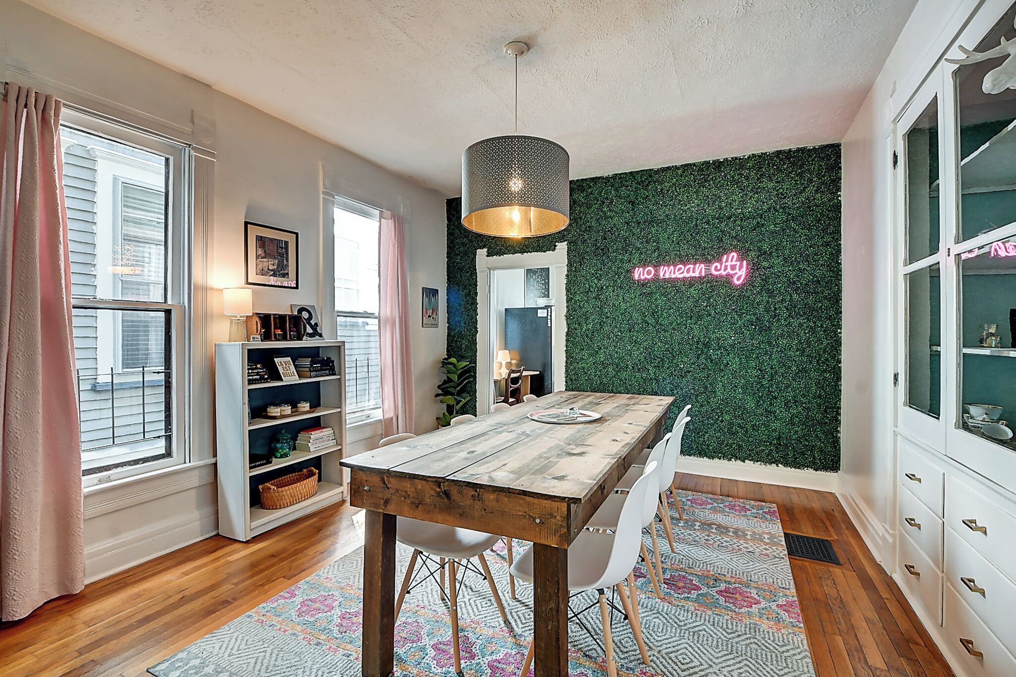 A dining area with a wooden table, white chairs, a hanging light fixture, a green wall with a neon sign, and a bookshelf.