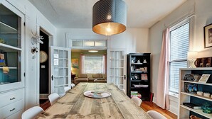 A dining room with a wooden table, white chairs, a chandelier, and a bookshelf. - Historic Talbott Street Home-Hot Tub and Fire Pit! (Indianapolis)