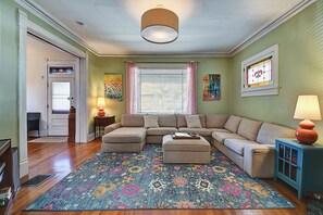 Un salón con un sofá modular, reposapiés, alfombra estampada y una ventana con persianas.