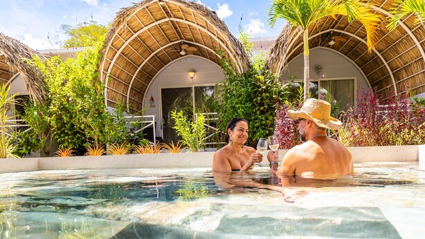 Una piscina con dos personas, rodeada de construcciones de techo de paja y abundante vegetación.