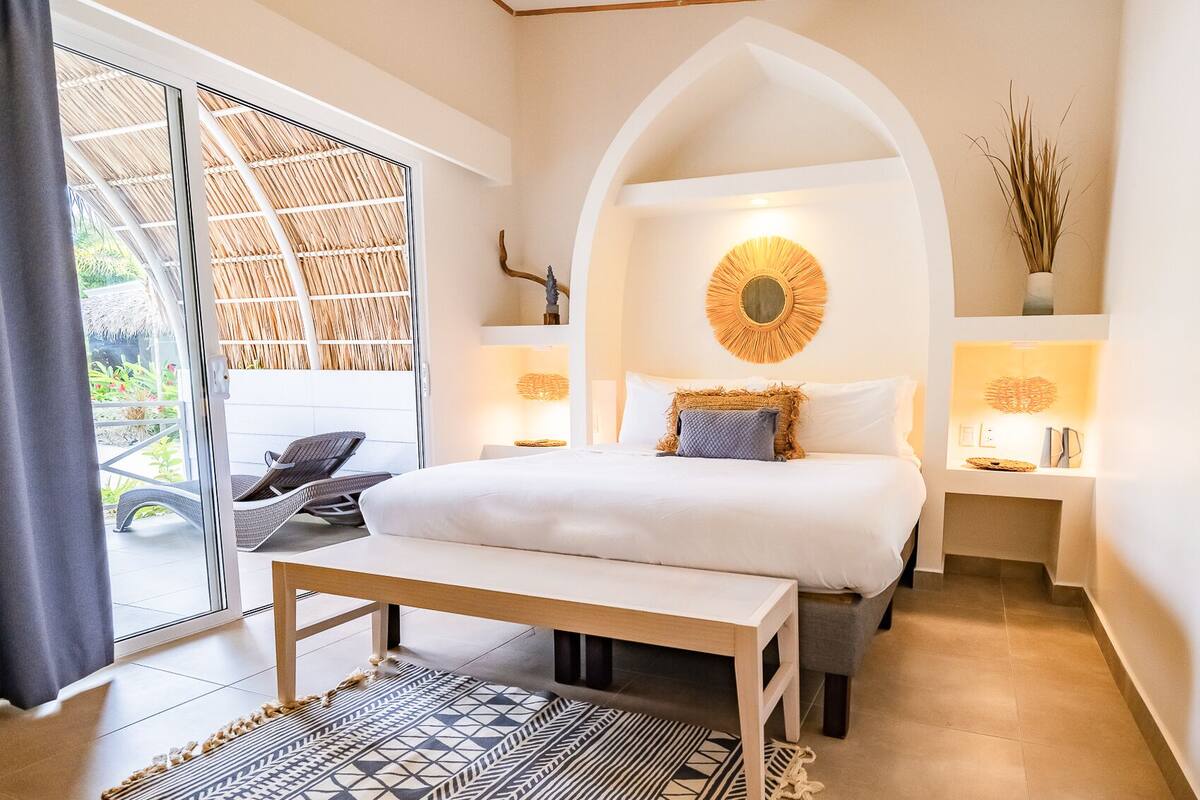 Une chambre à coucher avec un lit, un banc, un grand miroir et une vue sur l’extérieur.