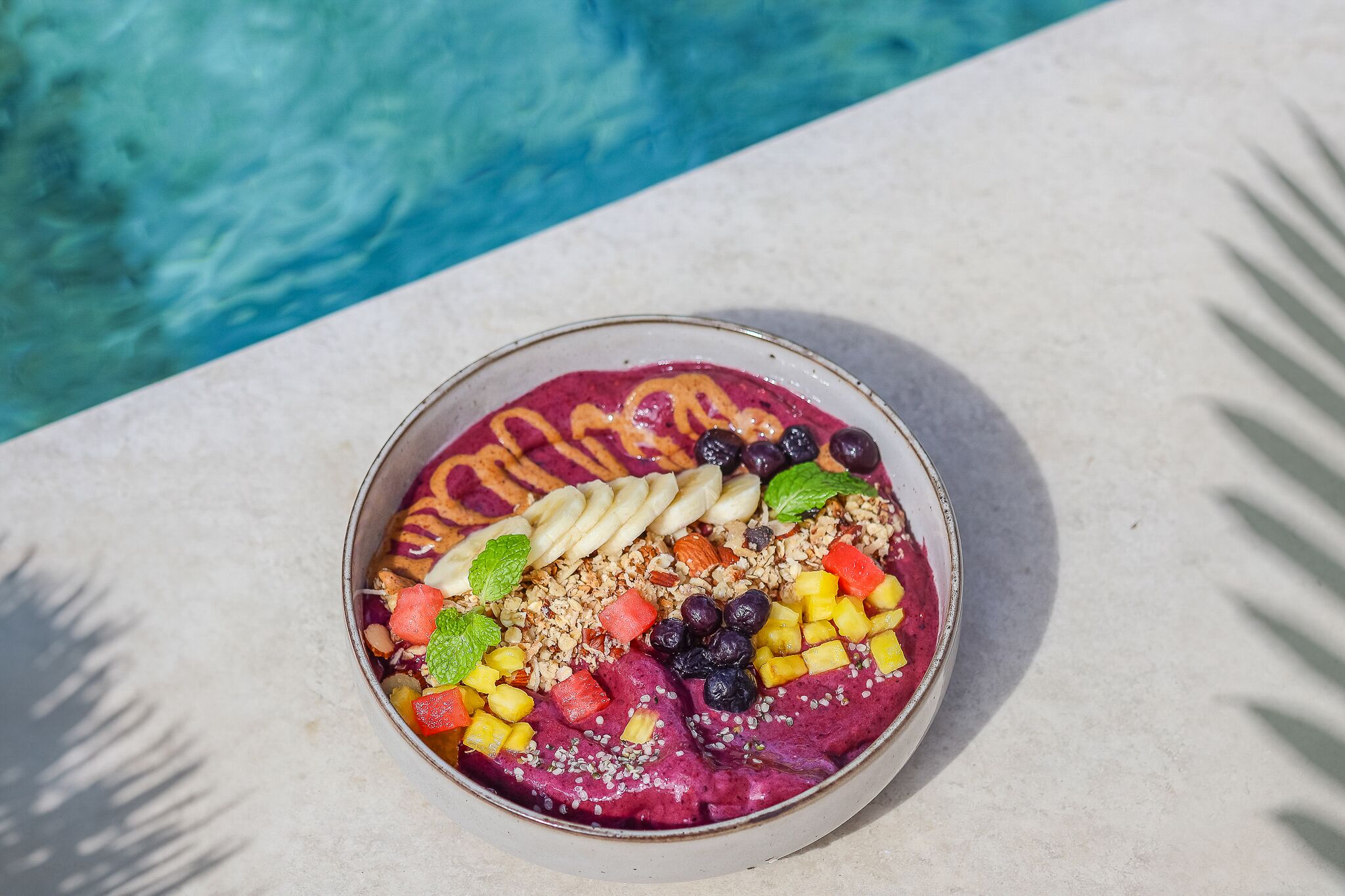 A bowl of smoothie bowl with various fruits and granola, placed on a table by a pool.
