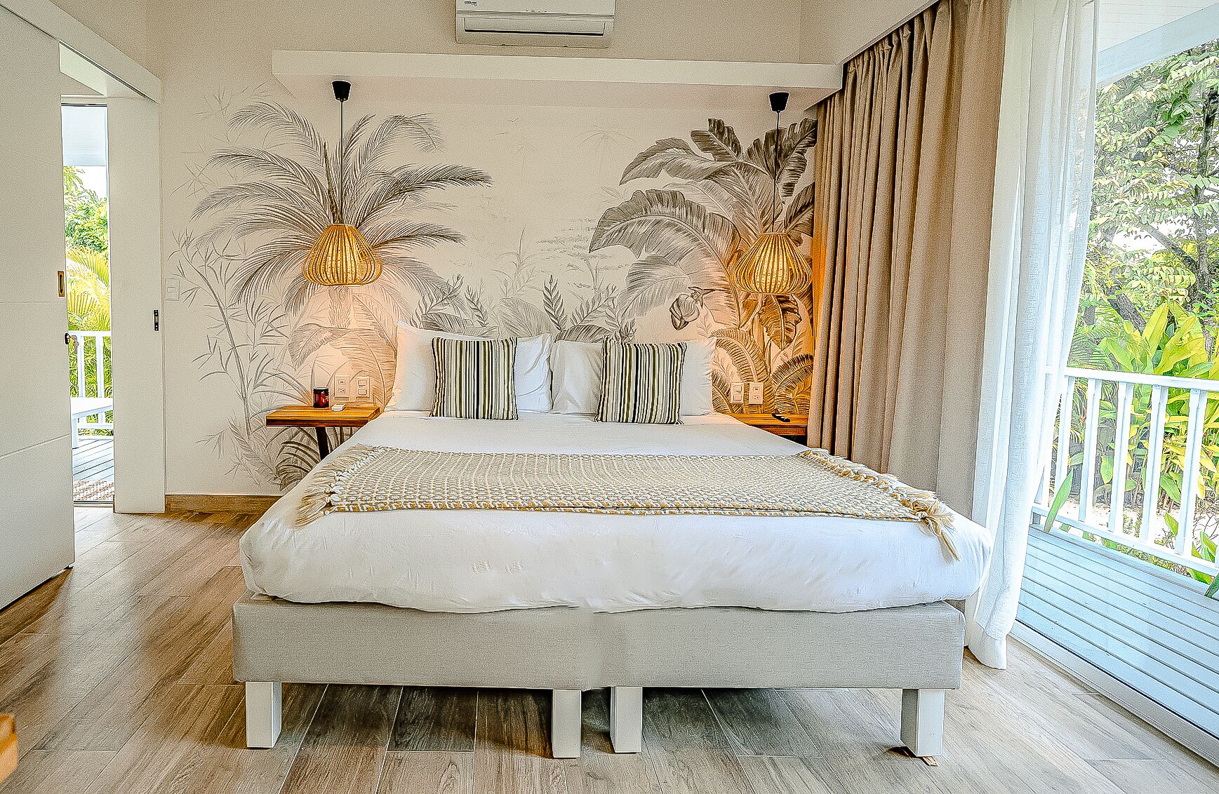 Een groot bed met een wit-beige beddekleed met dessin, twee nachtkastjes met lampen, en een muur met een tropische bladmuur.