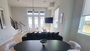Un salon moderne avec une grande table basse noire, un canapé et deux fauteuils blancs. Un escalier avec une rampe noire mène à un balcon.
