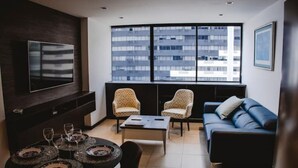A modern living room with a large window, a blue sofa, two patterned armchairs, a coffee table, and a dining table set with plates and glasses. - Puerto Santa Ana , automatic check in, Parking (Guayaquil)