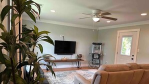 Ein Wohnzimmer mit einem beigen Sofa, einem Fernseher, einem Deckenventilator und einer großen Kübelpflanze.