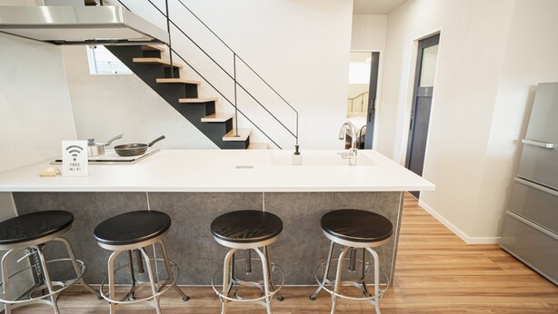 A modern kitchen with a white island, black stools, a stainless steel range hood, and a wooden staircase. - Orange County Guest House - Hostel  (Izumisano)