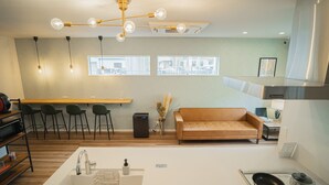 A modern kitchen with a large island, bar stools, a sofa, and a view of the city through large windows. - Orange County Guest House - Hostel  (Izumisano)