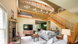 A spacious living room with a stone fireplace, a large grey sofa, a wooden coffee table, and a TV. A wooden staircase with a railing leads to an upper level. - Riverside Oasis! Close to Helen and Wineries (Suches)