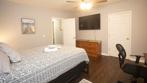 Un dormitorio con una cama, un aparador, una silla, un ventilador de techo y un televisor en la pared.