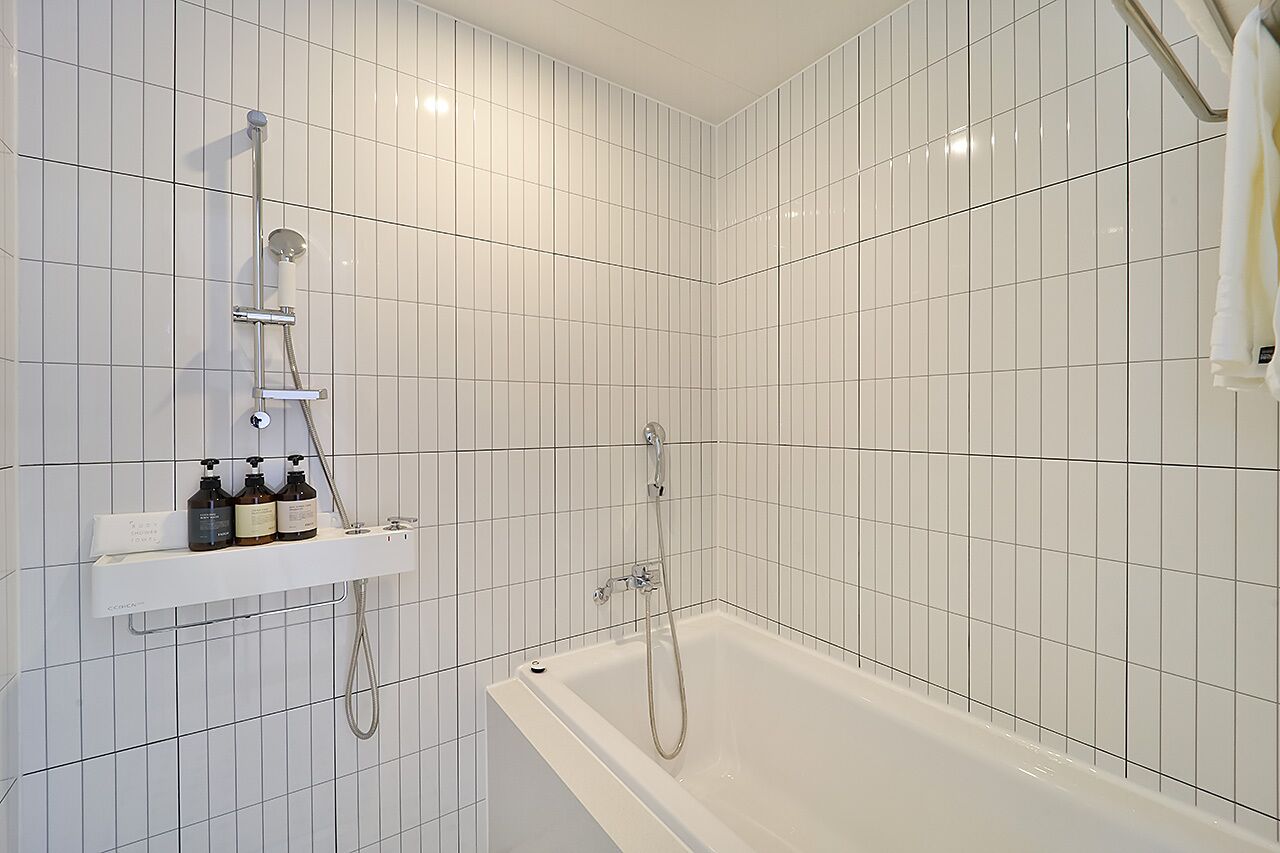 Une salle de bain moderne avec des murs recouverts de carreaux blancs, une baignoire, un espace douche et une étagère contenant des articles de toilette.