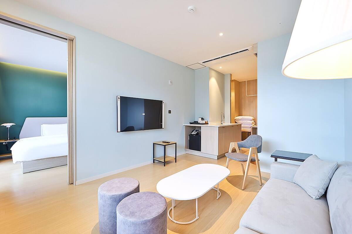 Moderni hotellihuone, jossa on sänky, televisio, pieni keittonurkkaus, ruokailutila ja sohva.