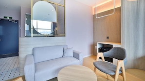 Una habitación de hotel moderna con sofá, una mesa redonda, una silla, televisor y un espejo grande.