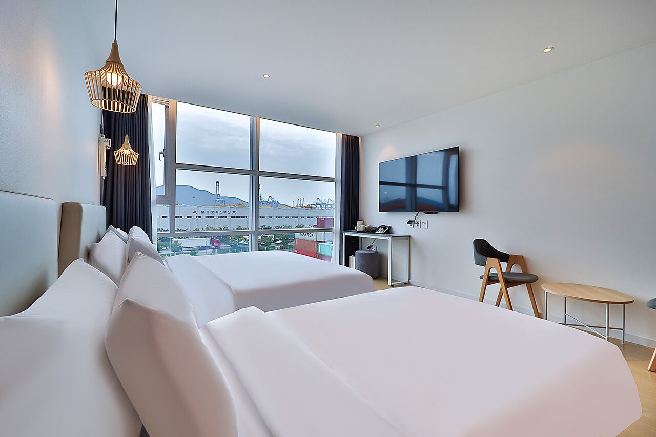 Une chambre d’hôtel avec deux lits, un bureau, une chaise, une télévision et une grande fenêtre donnant sur la ville.