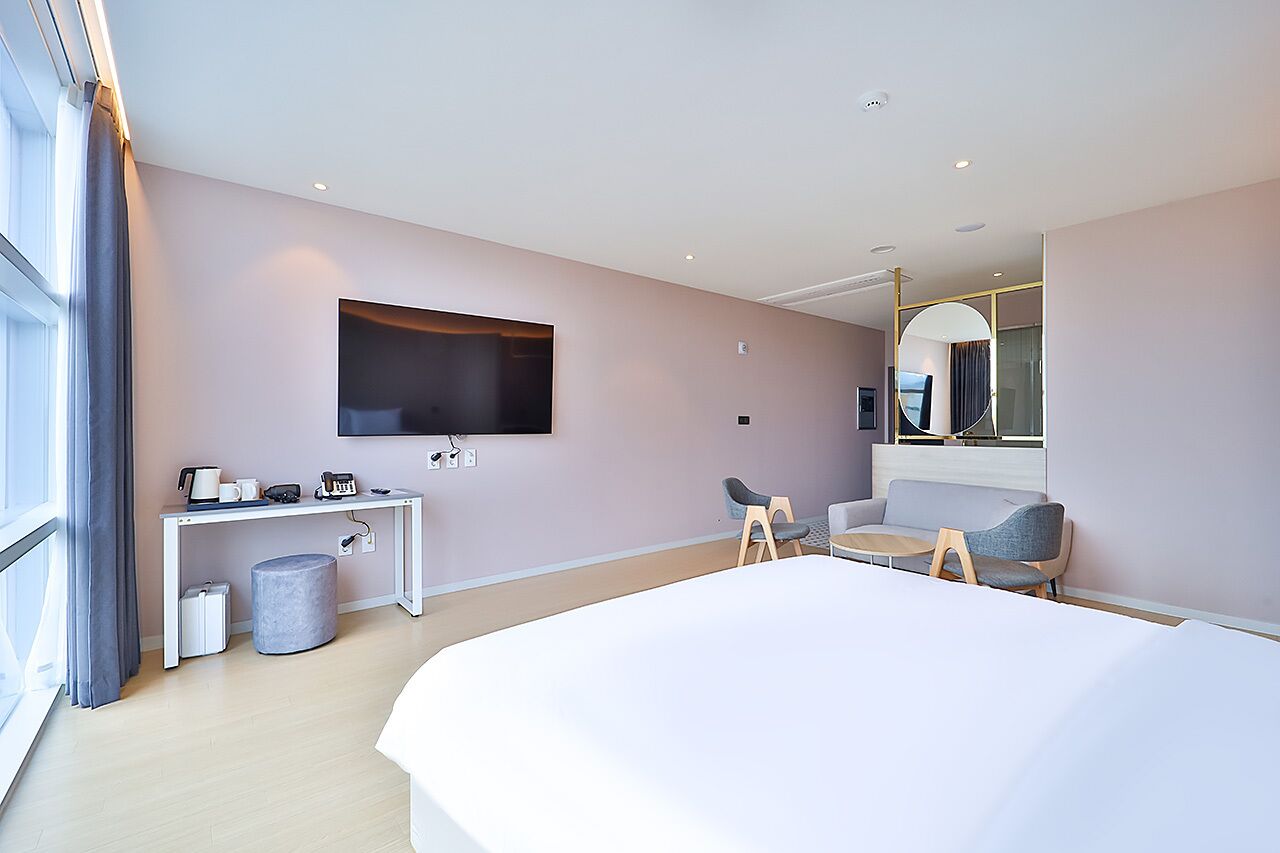 A modern hotel room with a large bed, a flat-screen TV mounted on the wall, a small desk with a chair, a round ottoman, and a sofa.