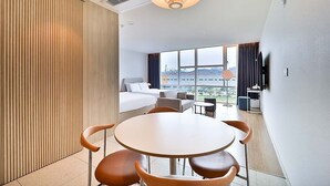 Habitación de hotel con cama, zona de comedor con mesa y sillas, televisor y ventana grande con vista a la ciudad.