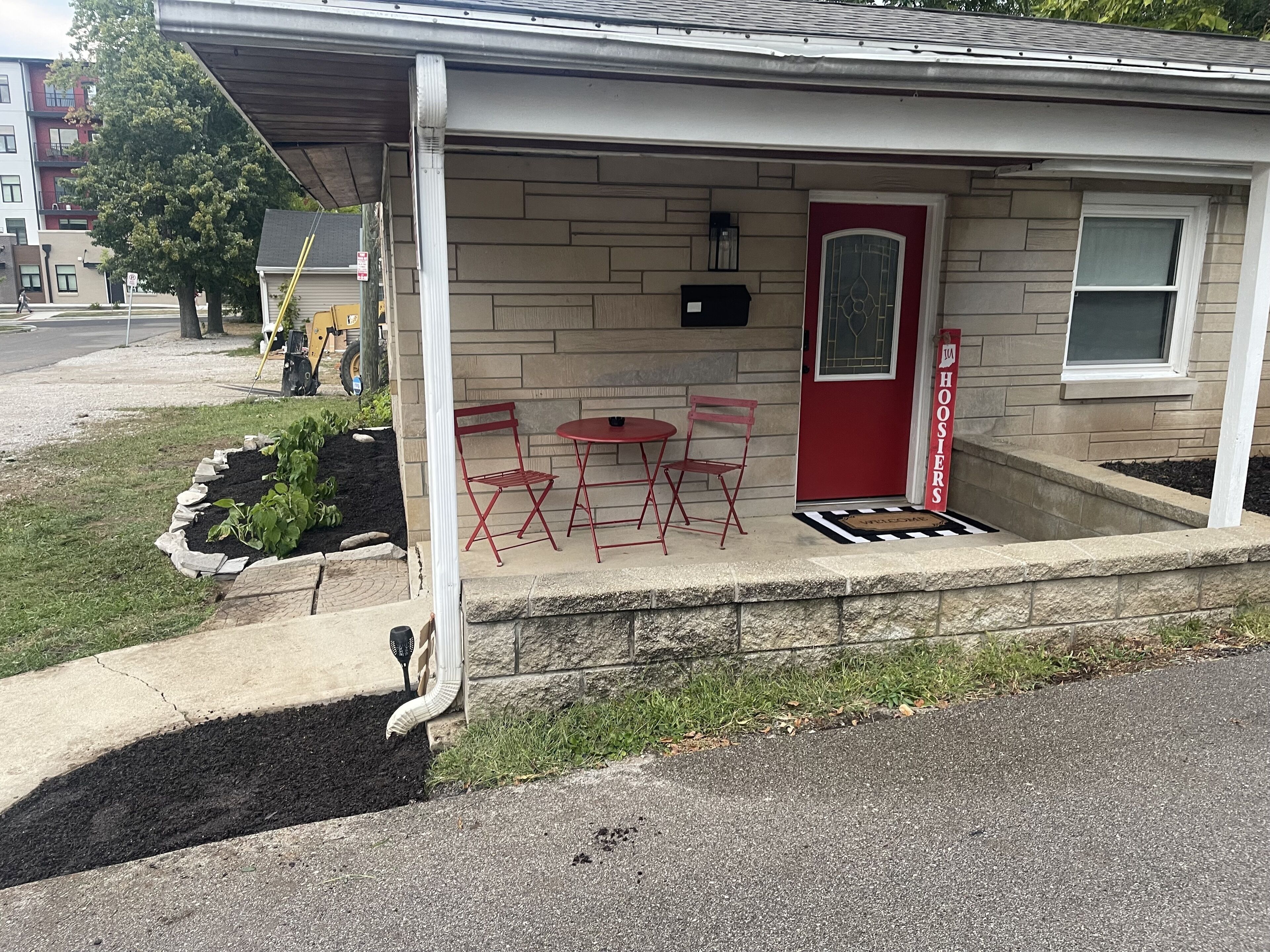 A small house with a red door, a porch with two red chairs and a table, and a welcome mat.