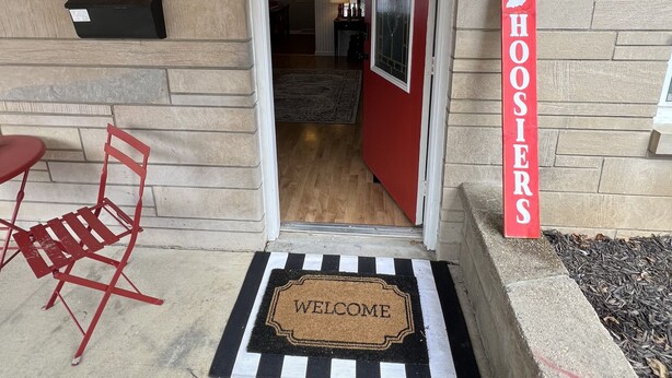 Una puerta roja con un felpudo que dice 'Welcome', un letrero rojo con 'HOOSIER'S' sobre una pared de piedra, y una silla plegable roja afuera.