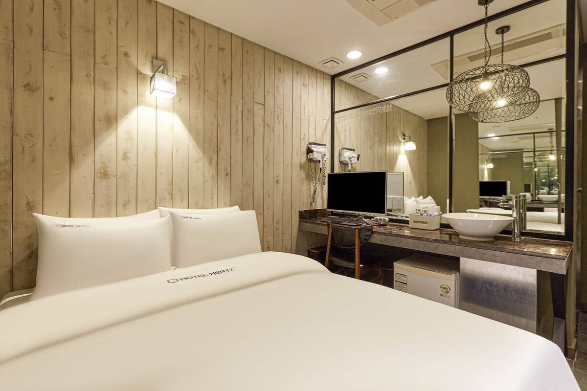 A hotel room with a wooden wall, a bed with white bedding, a desk with a computer, a sink, and a mirror.