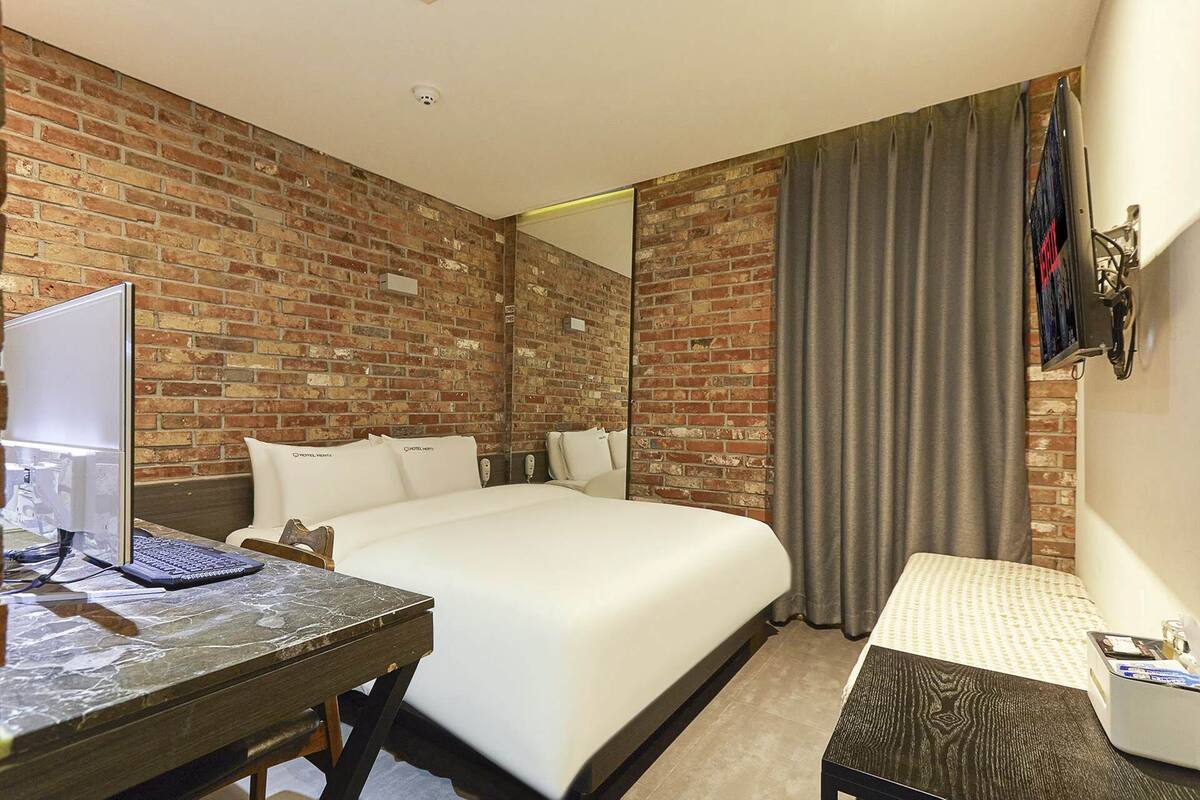 A hotel room with a brick wall, a bed, a desk with a computer, and a small table.