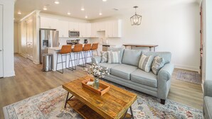 A modern kitchen with white cabinets, stainless steel appliances, and a dining area with a gray sofa and orange chairs.
