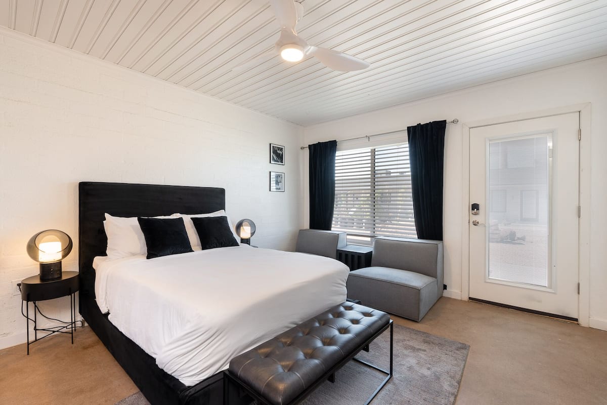 A bedroom with a large bed, a bench, a desk, and a window with curtains.