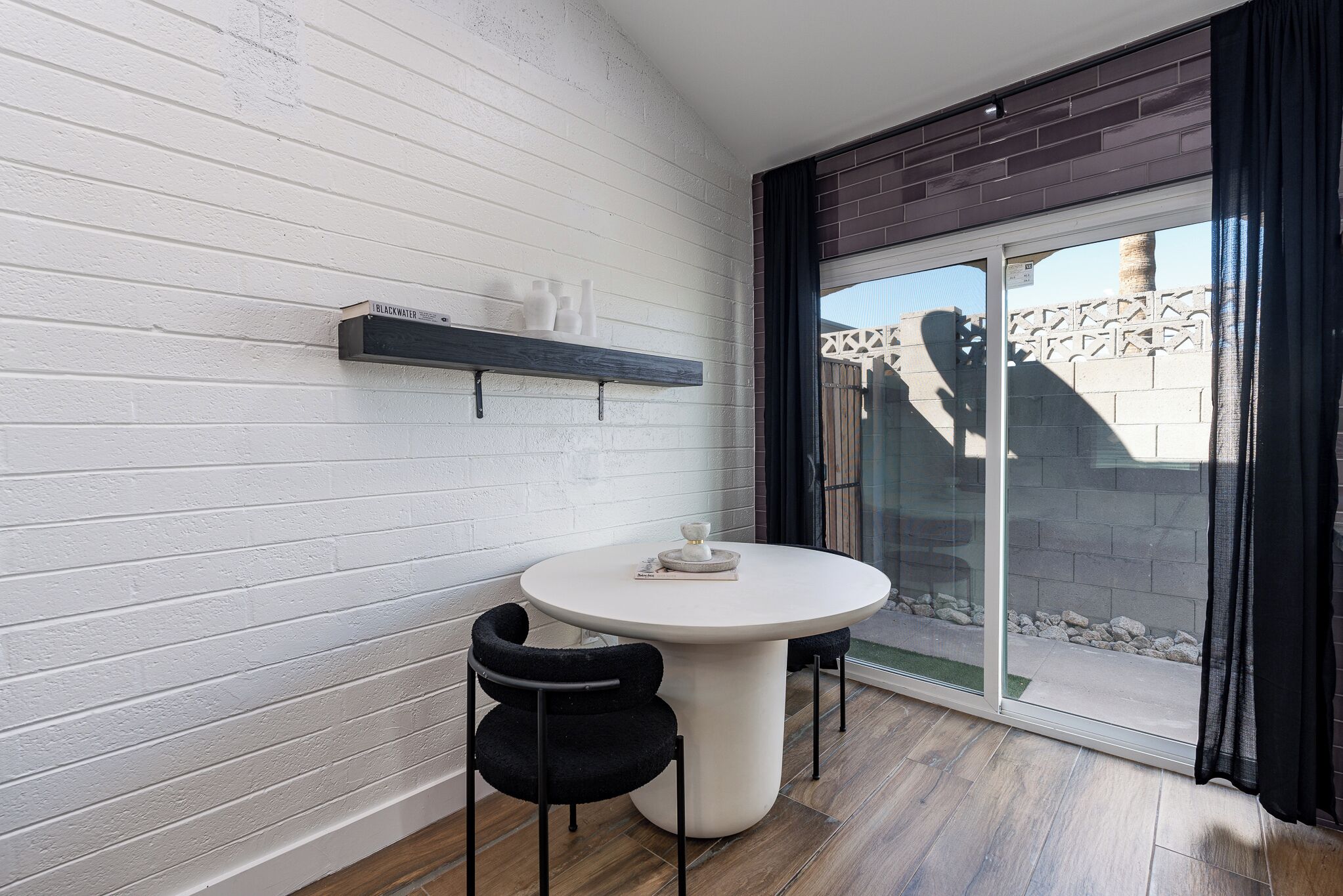 A small, round table with two black chairs, a white wall with horizontal lines, a dark brick accent wall, and a sliding glass door leading to an outdoor area.