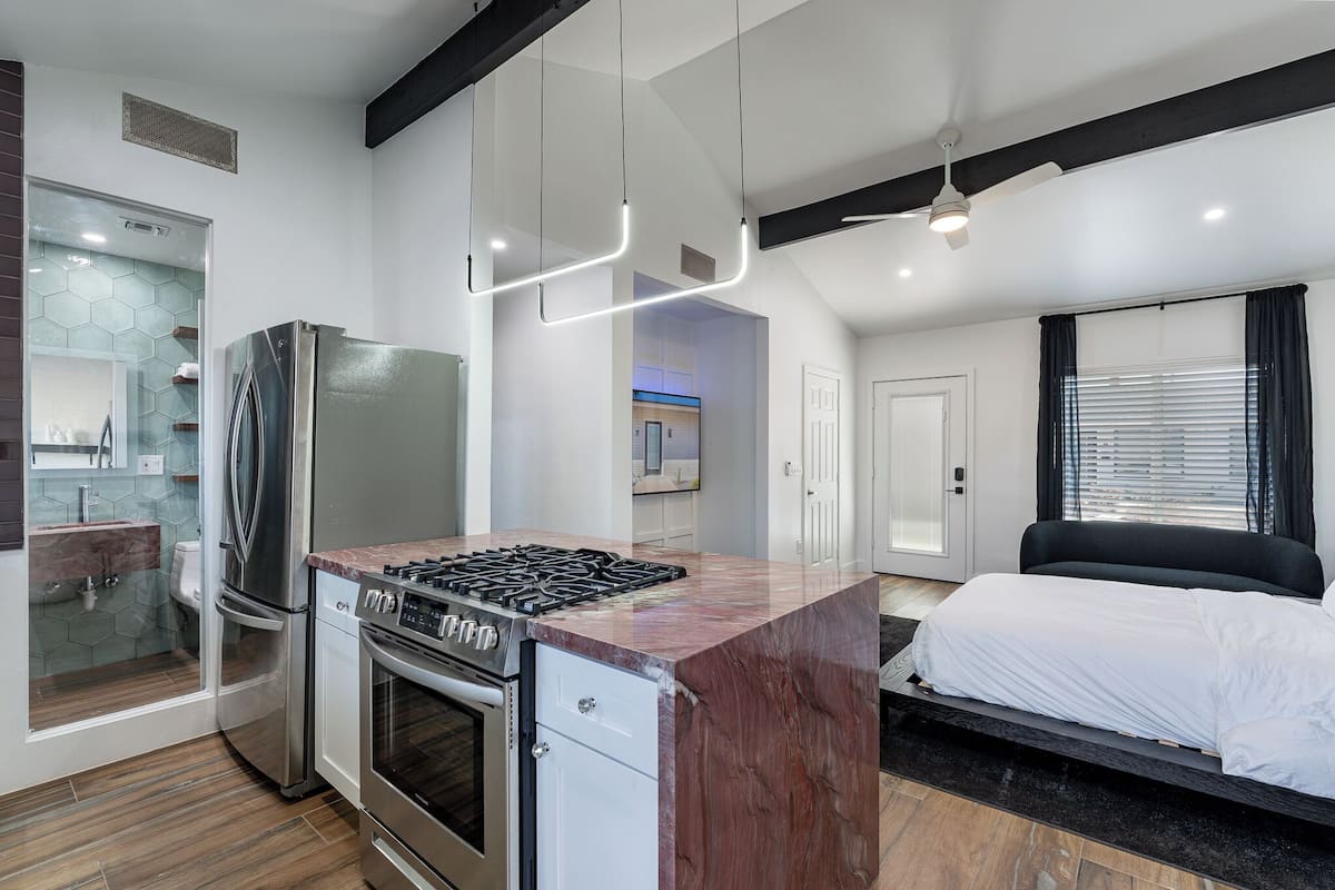 A modern kitchen with a built-in stove and refrigerator, a bedroom with a large bed and a window, and a bathroom with a glass shower enclosure.