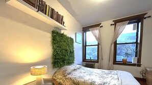 Een slaapkamer met een bed, een nachtkastje met een lamp, een boekenplank, een raam met gordijnen en een groene wanddecoratie.