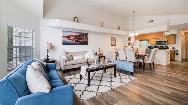 A spacious living room with a blue sofa, a beige sofa, and a dining area with a table and chairs. There is a kitchen in the background with wooden cabinets and a white ceiling fan. - Grand Superior Condo 3BD Apartment Disney Parks (Kissimmee)