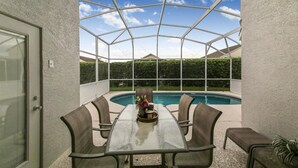 A covered patio with a glass table and chairs, a pool, and a view of a house and hedge. - Grand Luxury 4BD Pool Home Disney Universal (Davenport)