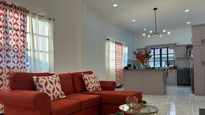 Ein geräumiges Wohnzimmer mit einem roten Sofa, einem Glastisch und einer Küchenzeile mit weißen Schränken und einer gefliesten Rückwand.