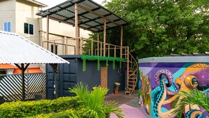 A building with a covered outdoor area, a mural with an octopus, and a garden with various plants. - Afueras Del Mar Container #1 (Lano Los Patos)