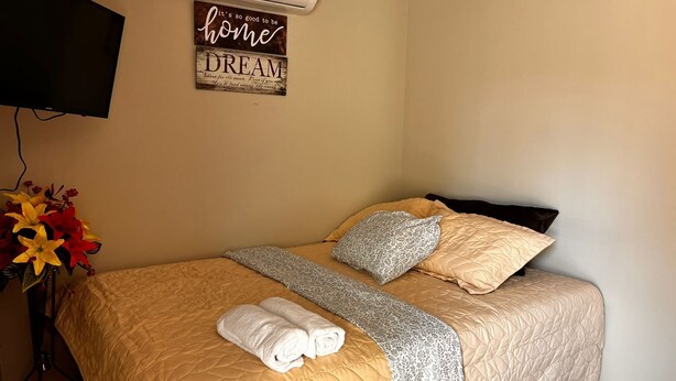 A neatly made bed with a quilted cover, two pillows, and a blanket. There is a wall-mounted air conditioner and a decorative sign above the bed. - Afueras Del Mar Container #1 (Lano Los Patos)