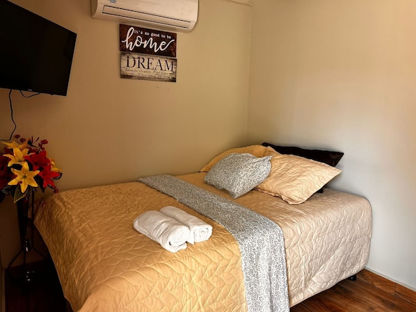A neatly made bed with a quilted cover, two pillows, and a blanket. There is a wall-mounted air conditioner and a decorative sign above the bed. - Afueras Del Mar Container #1 (Lano Los Patos)