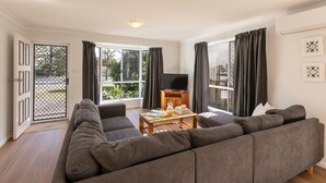 Een moderne woonkamer met een grote grijze modulaire bank, een houten salontafel en een flatscreen televisie op een houten standaard. Er zijn grote ramen met donkere gordijnen, een deur met een metalen hekwerk en een klein balkon is door de ramen zichtbaar.