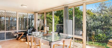 A spacious dining area with a glass table and chairs, large windows offering a view of the garden and trees.